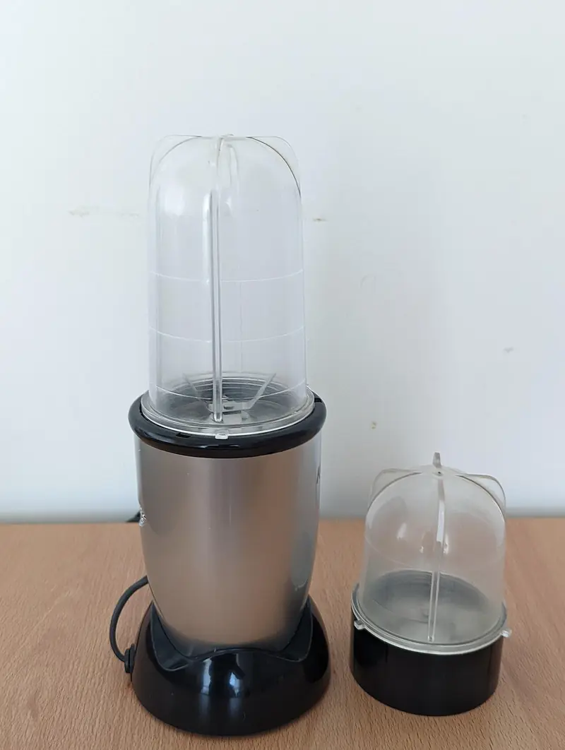 Blender mini berkabel dengan dudukan motor perak, gelas tinggi transparan, dan gelas kecil untuk bumbu di sampingnya